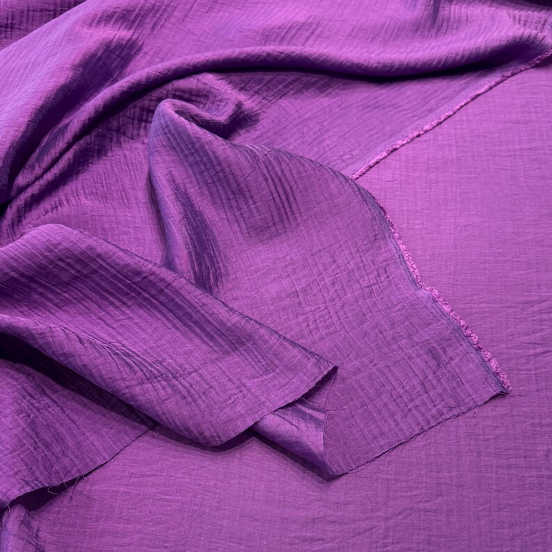 May include: A close-up of a vibrant purple fabric with a textured, crinkled appearance. The fabric is draped and folded, showcasing its flowing nature. The edges of the fabric are visible, revealing a raw, unfinished edge. The color is a rich, saturated purple.