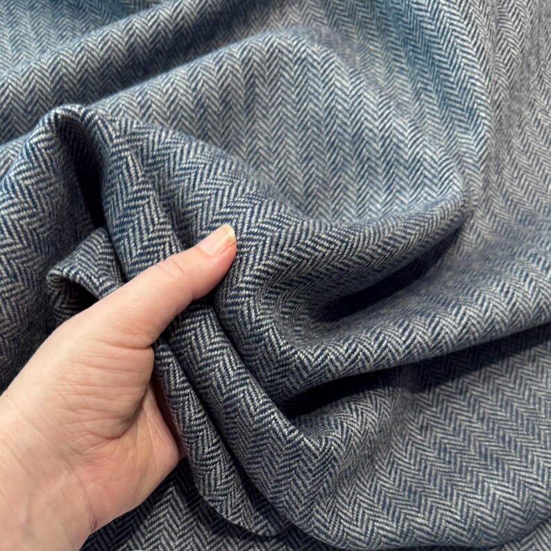 Eine Hand hält einen hochwertigen Fischgrät-Tweeds in Blau und Hellgrau, um die weiche, edle Qualität zu verdeutlichen. Der nachhaltig gefertigte Wollstoff aus Deutschland ist wärmend, langlebig und robust. Perfekt für Blazer, Jacken, leichte Mäntel.