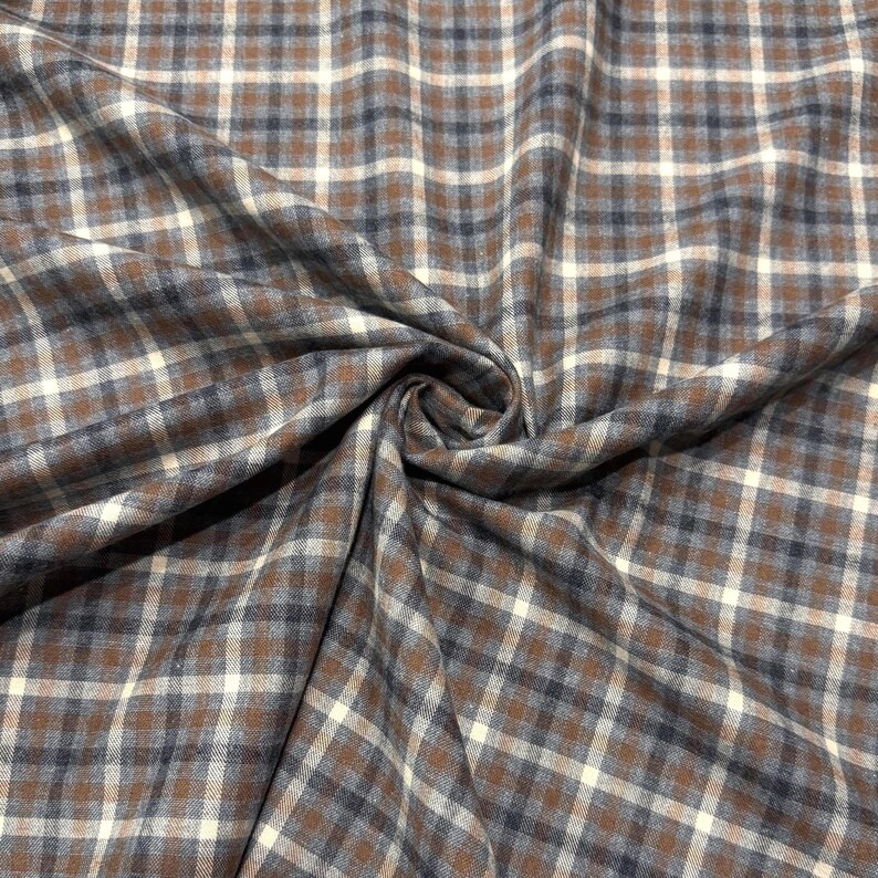 May include: Close-up of a fabric with a plaid pattern. The fabric features a repeating pattern of brown, white, and navy blue squares. The fabric is folded and draped, showing the texture and flow of the material.