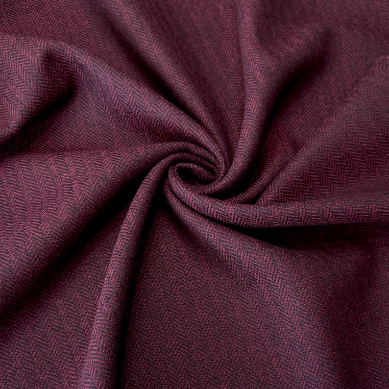 Detailaufnahme eines gefalteten Fischgrät-Tweeds in Weinrot und Schwarz. Die weiche Beschaffenheit und die exklusive Schurwolle machen den Stoff ideal für stilvolle Bekleidungsstücke wie Mäntel, Capes und winterwarme Westen.