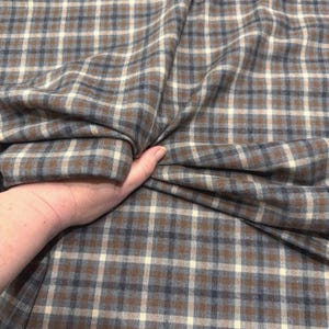 May include: A close-up of a fabric with a plaid pattern in shades of brown, blue, and cream. The fabric is being held in a hand, showing the texture and drape of the material. The plaid design is a classic pattern.