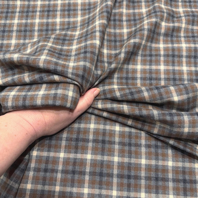 May include: A close-up of a fabric with a plaid pattern in shades of brown, blue, and cream. The fabric is being held in a hand, showing the texture and drape of the material. The plaid design is a classic pattern.