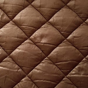 Könnte beinhalten: Nahaufnahme eines braunen, gesteppten Stoffes mit Rautenmuster. Der Stoff hat ein leicht glänzendes Aussehen und eine strukturierte Oberfläche. Die Steppung erzeugt ein Gitter aus Rautenformen.