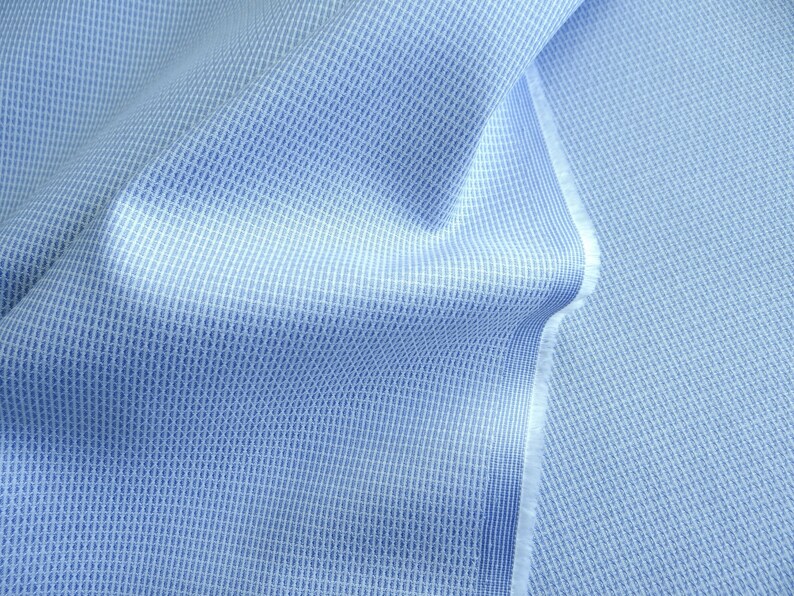 May include: Close-up of a light blue fabric with a textured, woven pattern. The fabric has a subtle grid-like design, with a lighter shade of blue creating a contrast. The edges of the fabric are visible, showing the raw, unfinished edge.