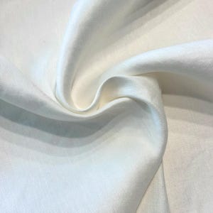 May include: Close-up of white fabric with gentle folds and a subtle texture. The material looks lightweight, possibly linen or a similar textile, with a smooth, slightly shiny surface. The folds create shadows and highlights.