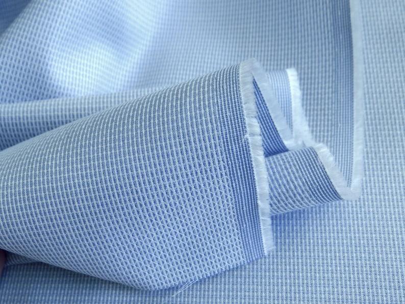 May include: Close-up of a light blue fabric with a textured weave. The fabric has a grid-like pattern with vertical stripes and a subtle, raised texture. The edges of the fabric are frayed, revealing the raw, unfinished edges of the material.