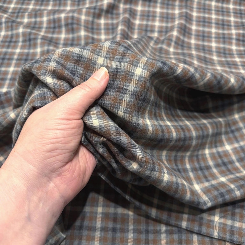 May include: A close-up of a fabric with a plaid pattern. The fabric is a mix of brown, blue, and off-white squares. The fabric is being held in a hand, showing the texture and drape of the material. The fabric appears to be a medium weight.
