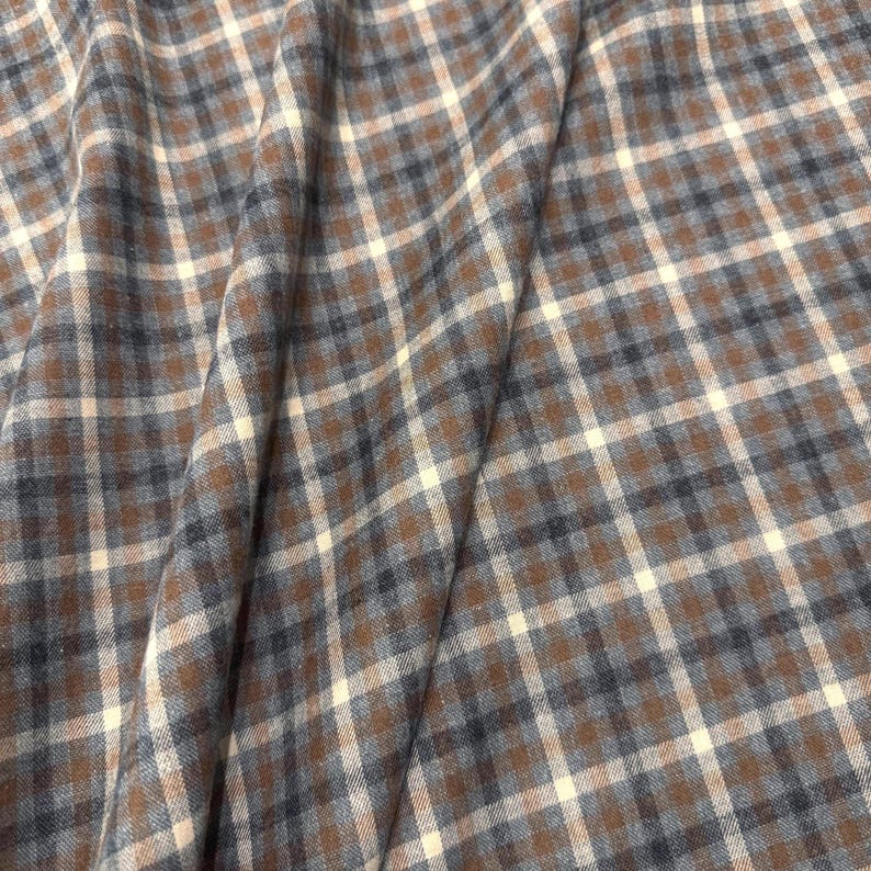 May include: Close-up of a fabric with a plaid pattern. The fabric features a repeating pattern of brown, off-white, and navy blue squares. The fabric is folded, creating shadows and depth, and is likely used for clothing or home decor.