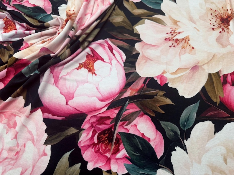 Pode incluir: Close-up de tecido com um padr&atilde;o floral. Grandes pe&oacute;nias cor-de-rosa e folhas verde-escuras num fundo preto. O tecido est&aacute; dobrado, mostrando a textura e profundidade do design.