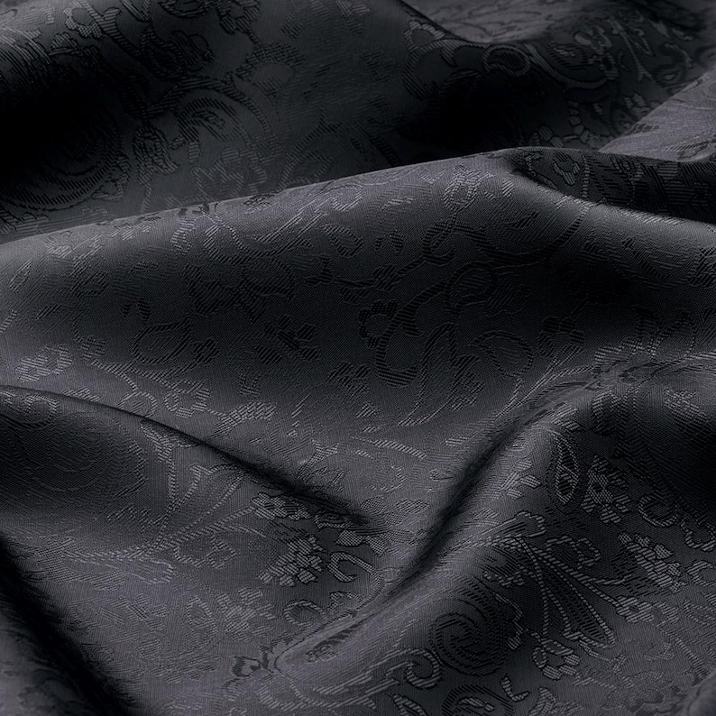 May include: A black fabric featuring a tonal jacquard weave with an intricate floral and paisley pattern. The material displays a subtle sheen and is captured in soft, flowing folds that highlight the depth and texture of the woven design.