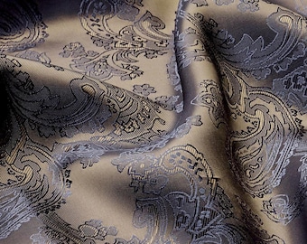May include: A fabric with a woven paisley pattern featuring dark blue and charcoal grey motifs set against a shimmering light bronze background. The material displays deep folds and creases that highlight the intricate texture and sheen.