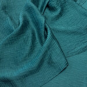 May include: Close-up of teal fabric with a textured, crinkled appearance. The material is folded, creating shadows and highlighting the fabric's sheen. The color is a deep, saturated teal, suggesting a luxurious textile.