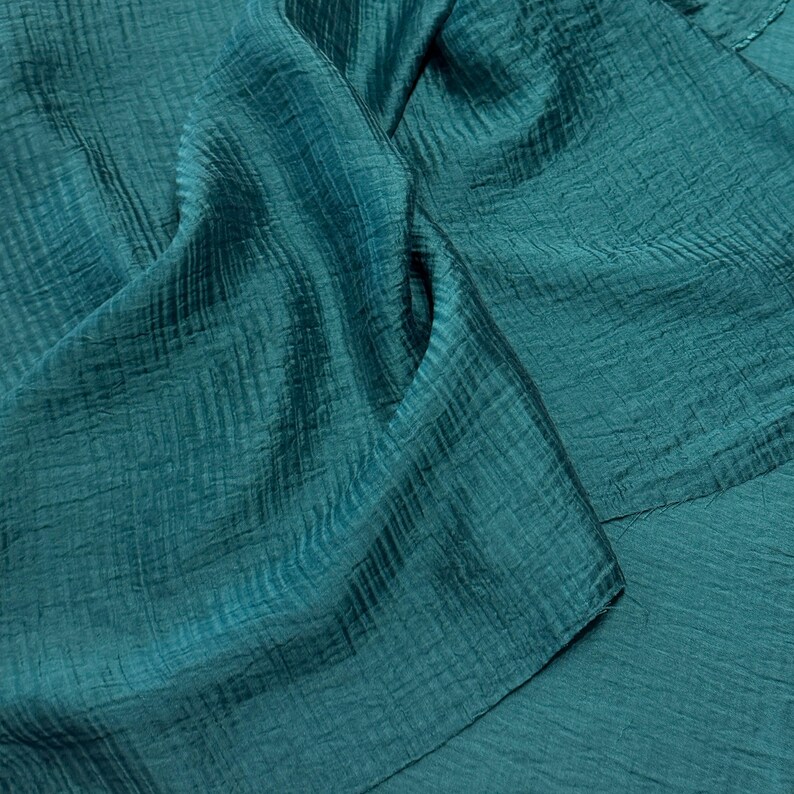 May include: Close-up of teal fabric with a textured, crinkled appearance. The material is folded, creating shadows and highlighting the fabric's sheen. The color is a deep, saturated teal, suggesting a luxurious textile.