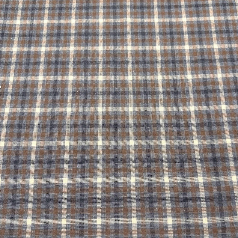 May include: A close-up of a fabric with a plaid pattern. The pattern features a grid of intersecting lines in brown, white, and navy blue. The fabric has a textured appearance.