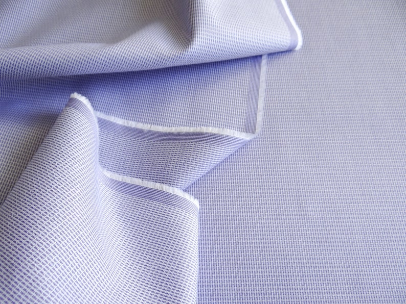 May include: Close-up of a light blue woven fabric with a textured pattern. The fabric is folded, revealing a white edge. The material has a subtle grid-like design, suitable for apparel or home decor.