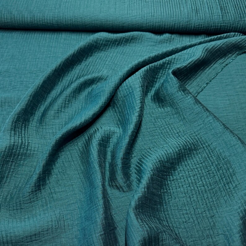 May include: Teal-colored fabric with a textured, crinkled appearance. The fabric is draped, showing folds and shadows that highlight the texture. The material has a slight sheen, suggesting it could be silk or a similar fabric.
