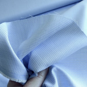 May include: Close-up of light blue fabric with a textured weave and white stripes. The fabric is folded, revealing a solid light blue material underneath. The edges of the fabric are frayed.