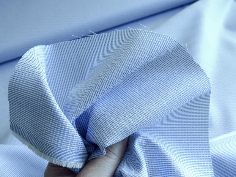 May include: Close-up of light blue fabric with a textured weave and white stripes. The fabric is folded, revealing a solid light blue material underneath. The edges of the fabric are frayed.