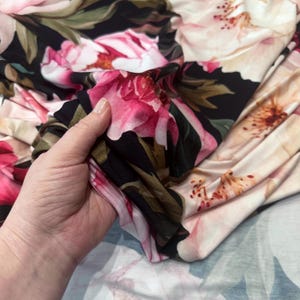 Pode incluir: Tecido floral com grandes flores rosa e creme sobre um fundo preto. O tecido &eacute; segurado em uma m&atilde;o, mostrando a estampa detalhada e a textura macia. Folhas verdes e detalhes completam o design.