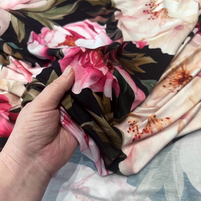Pode incluir: Tecido floral com grandes flores rosa e creme sobre um fundo preto. O tecido &eacute; segurado em uma m&atilde;o, mostrando a estampa detalhada e a textura macia. Folhas verdes e detalhes completam o design.