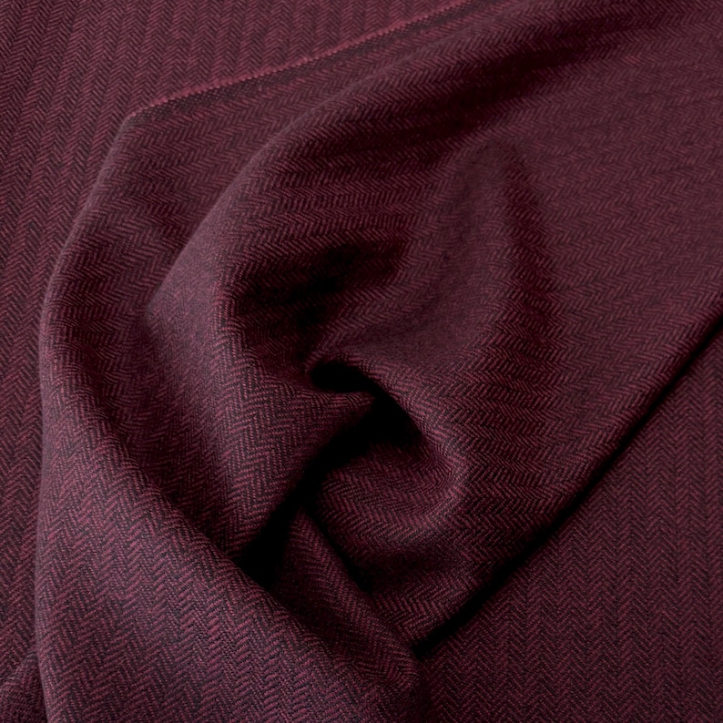 Detailaufnahme eines gefalteten Fischgrät-Tweeds in Weinrot und Schwarz. Die weiche, wollige Textur und das klassische Muster sind klar zu erkennen. Der Stoff eignet sich hervorragend für winterwarme Blazer, leichte Mäntel und stilvolle Accessoires.