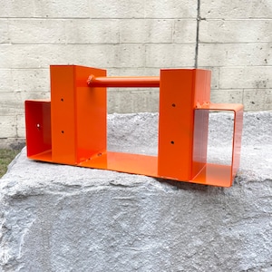 Puede incluir: Marco de metal naranja con una barra horizontal, sentado sobre un bloque de hormigón gris. El marco tiene varios agujeros y está diseñado para montaje.