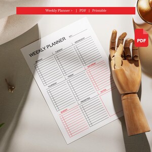May include: A white weekly planner with a red header and accents. The planner includes sections for each day of the week, priorities, and notes. The text "Weekly Planner" is at the top. A wooden hand prop is next to the planner.