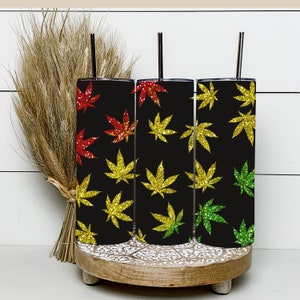 May include: Three stainless steel tumblers with black backgrounds and glitter marijuana leaf designs. The leaves are red, yellow, and green.
