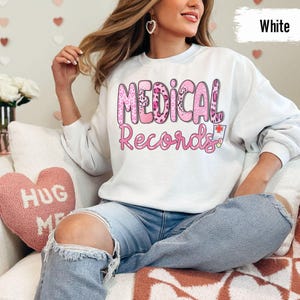 May include: White sweatshirt with the text "MEDICAL RECORDS" in pink letters with a leopard print design. The sweatshirt is worn by a person sitting on a couch with a pink heart-shaped pillow that says "HUG ME".