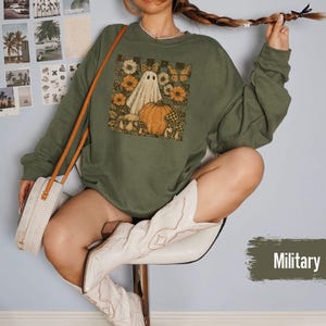 May include: An olive green sweatshirt featuring a graphic of a ghost, pumpkin, flowers, and butterflies. The sweatshirt is long-sleeved and has a crew neck. The person in the image is wearing white cowboy boots and carrying a white purse.
