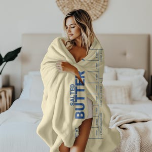 May include: A cream-colored throw blanket with the words "NET WT. SALTED BUTTER" printed in blue on the side. The blanket has a measurement guide printed on the side in blue, with markings for tablespoons and cups.