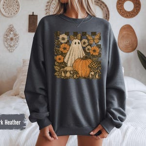 May include: Dark heather gray sweatshirt featuring a vintage-style graphic of a ghost, pumpkin, flowers, and mushrooms. The design includes a checkered background and butterflies, evoking a fall or Halloween theme.