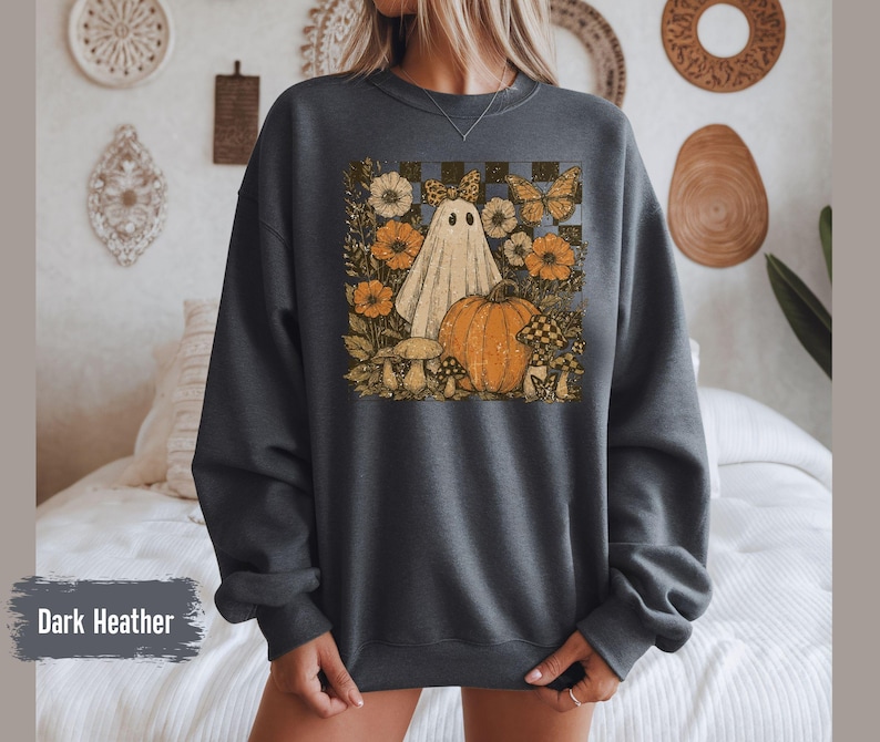 May include: Dark heather gray sweatshirt featuring a vintage-style graphic of a ghost, pumpkin, flowers, and mushrooms. The design includes a checkered background and butterflies, evoking a fall or Halloween theme.