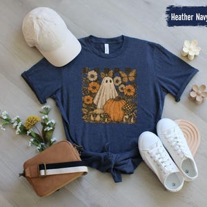 May include: Heather navy t-shirt with a whimsical Halloween graphic. The design features a ghost, pumpkin, flowers, and butterflies. A white baseball cap, brown purse, and white sneakers are also in the image. The text "Heather Navy" is visible.