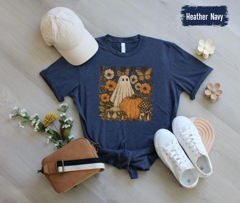 May include: Heather navy t-shirt with a whimsical Halloween graphic. The design features a ghost, pumpkin, flowers, and butterflies. A white baseball cap, brown purse, and white sneakers are also in the image. The text "Heather Navy" is visible.