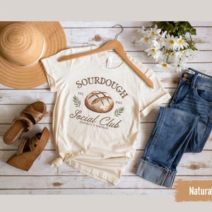 May include: A beige t-shirt with a brown sourdough bread graphic and the text "Sourdough Social Club Est. 2023 Happiness is Homemade". The shirt is laid out on a white wooden surface with a straw hat, brown sandals, a bouquet of white flowers, and blue jeans.