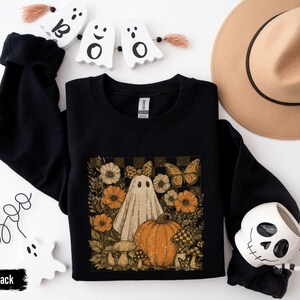May include: Black crewneck sweatshirt featuring a vintage-style Halloween graphic. The design includes a ghost, pumpkin, flowers, and butterflies. The word "Boo" is written in cursive, and the word "Black" is in a black rectangle.