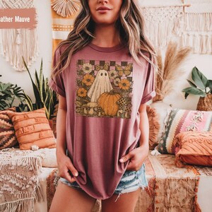 May include: Heather mauve t-shirt with a graphic featuring a ghost, pumpkin, flowers, and butterflies. The design is set against a checkered background. The shirt has short sleeves and a relaxed fit.