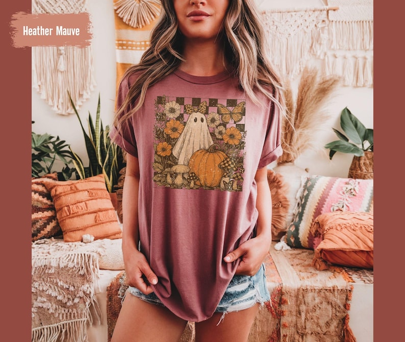 May include: Heather mauve t-shirt with a graphic featuring a ghost, pumpkin, flowers, and butterflies. The design is set against a checkered background. The shirt has short sleeves and a relaxed fit.