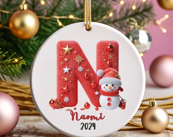 May include: Red and white Christmas bauble featuring a large letter N, adorned with stars and snowflakes. A snowman wearing a red hat and scarf is positioned in front of the letter. The bauble is personalised with the name Naomi and the year 2024.