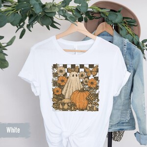 May include: White t-shirt featuring a Halloween-themed graphic. The design includes a ghost, pumpkin, flowers, and butterflies set against a checkered background. The shirt is hanging on a wooden hanger.