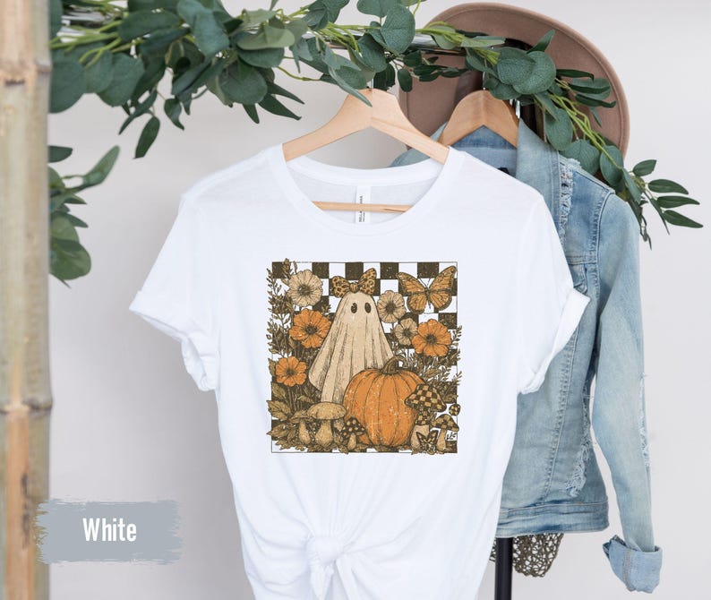 May include: White t-shirt featuring a Halloween-themed graphic. The design includes a ghost, pumpkin, flowers, and butterflies set against a checkered background. The shirt is hanging on a wooden hanger.