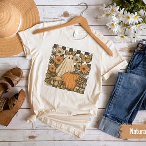 May include: Cream-colored t-shirt featuring a Halloween-themed graphic with a ghost, pumpkin, and flowers. The shirt is displayed with a straw hat, brown sandals, and blue jeans on a wooden surface. The word "Natural" is visible.
