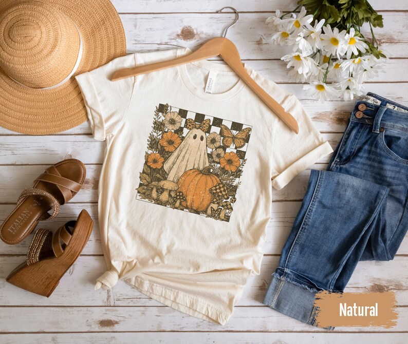 May include: Cream-colored t-shirt featuring a Halloween-themed graphic with a ghost, pumpkin, and flowers. The shirt is displayed with a straw hat, brown sandals, and blue jeans on a wooden surface. The word "Natural" is visible.