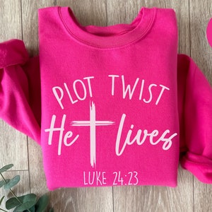 May include: Pink sweatshirt with the text "PLOT TWIST He lives" and "LUKE 24:23" in white.