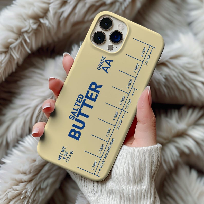 May include: A beige iPhone case with a stick of butter design. The case features a blue label with the text "SALTED BUTTER AA" and a measurement guide in tablespoons and cups. The case has a clear plastic window for the phone camera.
