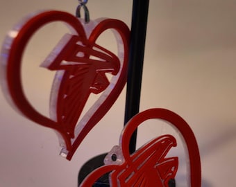 May include: A pair of red acrylic heart-shaped earrings featuring the Atlanta Falcons logo in the center. The earrings are suspended from silver-toned metal hooks and display a layered design with a solid red outline and a cutout falcon emblem.