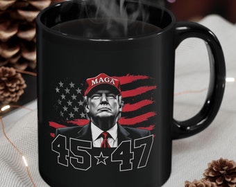 May include: Black coffee mug featuring a red, white and blue American flag design. The flag has a white star in the blue field. The mug has a white outline of the numbers 45 and 47 with a star between the numbers. The mug also features the text "MAGA" in red letters above a portrait of a man wearing a red "MAGA" hat.