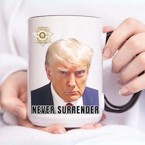 May include: White ceramic mug with a black handle. The mug features a photo of Donald Trump and the text "NEVER SURRENDER". A gold sheriff's star badge is visible in the background.