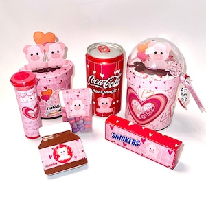 Könnte beinhalten: Eine Sammlung rosa Valentinstagsgeschenke, darunter eine Coca-Cola-Dose, ein Nutella-Glas, ein Snickers-Riegel und verschiedene Schachteln, alle mit Schweineillustrationen und Herzmotiven verziert.